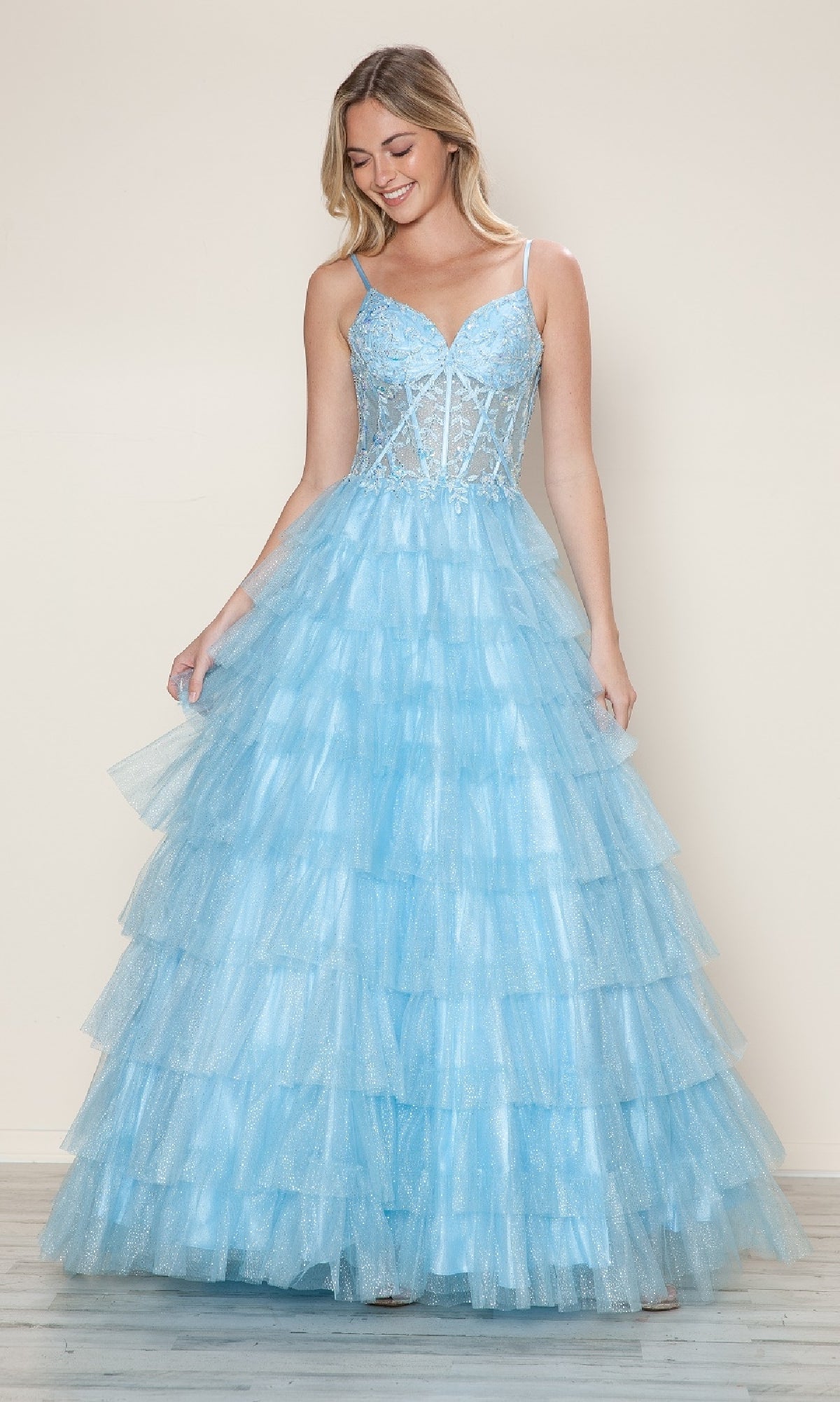 A woman wears the Long Prom Dress: Poly USA 9608—a light blue, ruffled ball gown with a tiered skirt, thin straps, and a semi-sheer lace corset bodice—standing against a plain beige background for an ethereal look.